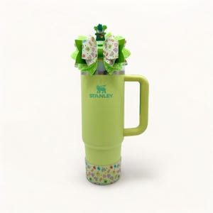 May include: A green Stanley travel mug with a green, white, and gold shamrock bow on top. The mug has a handle and the word "STANLEY" is printed on the side.