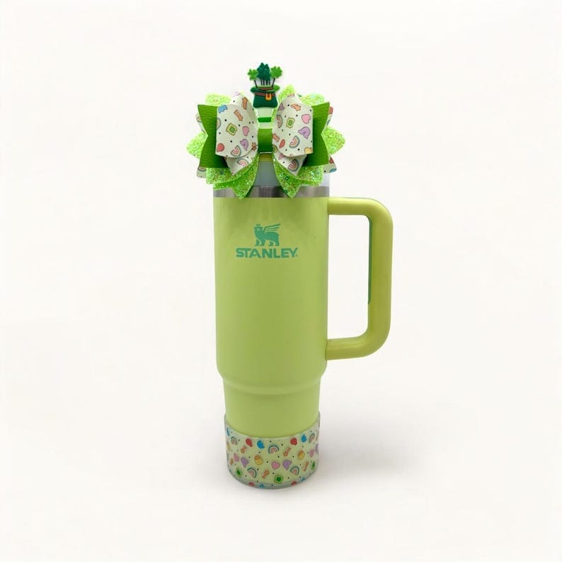 May include: A green Stanley travel mug with a green, white, and gold shamrock bow on top. The mug has a handle and the word "STANLEY" is printed on the side.