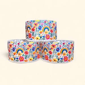 May include: Three clear glass candle holders with a colorful floral and rainbow pattern. The pattern features a white background with multi-colored flowers, leaves, and rainbows.