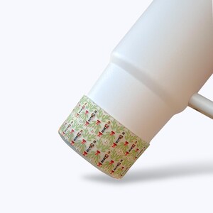 May include: A white tumbler with a decorative band featuring a festive pattern. The band showcases green pine branches, red bows, and black and white elements, creating a holiday theme. The tumbler has a white handle.