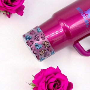 May include: A pink Stanley thermos with a leopard print heart design on the lid. The thermos has a handle and a silver logo that says "Stanley".