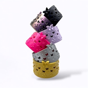 May include: A stack of five colorful, cylindrical bracelets. Each bracelet is a different color: gold, silver, pink, lavender, and black. The bracelets are decorated with butterfly cutouts and raised butterfly accents.