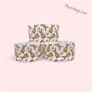 May include: Three white fabric rolls with a repeating pattern of brown bunnies wearing pink bows. The text "Matching bow" is written in pink at the top of the image.