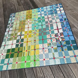 May include: A square patchwork quilt featuring a grid of multi-colored fabric squares in shades of green, yellow, teal, and blue. The design includes small red square accents at the intersections of the grid, with visible swirling quilted stitching.