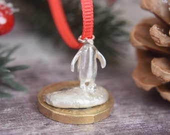 May include: A silver penguin ornament with a red ribbon for hanging. The penguin stands on a small, round, silver base atop a gold coin. The background includes red berries and pine needles, suggesting a holiday theme.
