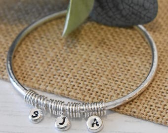 May include: A silver-toned bangle bracelet featuring a hammered texture and a series of metallic coils. Three small, round, silver-toned charms are suspended from the coils, each stamped with a single black letter: S, J, and A respectively.