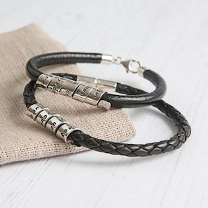 May include: Two black leather bracelets with silver beads. The beads are engraved with letters. The bracelets are on a light brown fabric surface.