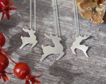 May include: Three silver-toned reindeer pendant necklaces. Each pendant is a cutout of a reindeer silhouette. The necklaces hang on delicate silver chains. The background is a rustic wooden surface with red berries and a pine cone.