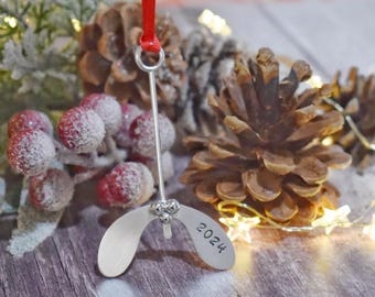 May include: A silver-colored metal Christmas ornament shaped like mistletoe, with the year 2024 engraved on one of the leaves. It hangs from a red ribbon. The background includes pine cones, frosted red berries, and fairy lights.