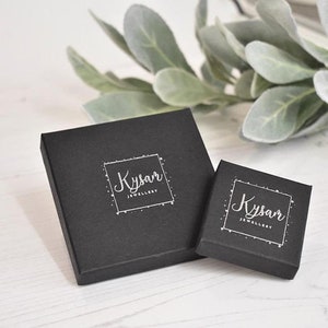 May include: Two black square boxes with the text "Kysar Jewellery" in white lettering. The boxes are on a white wooden surface.