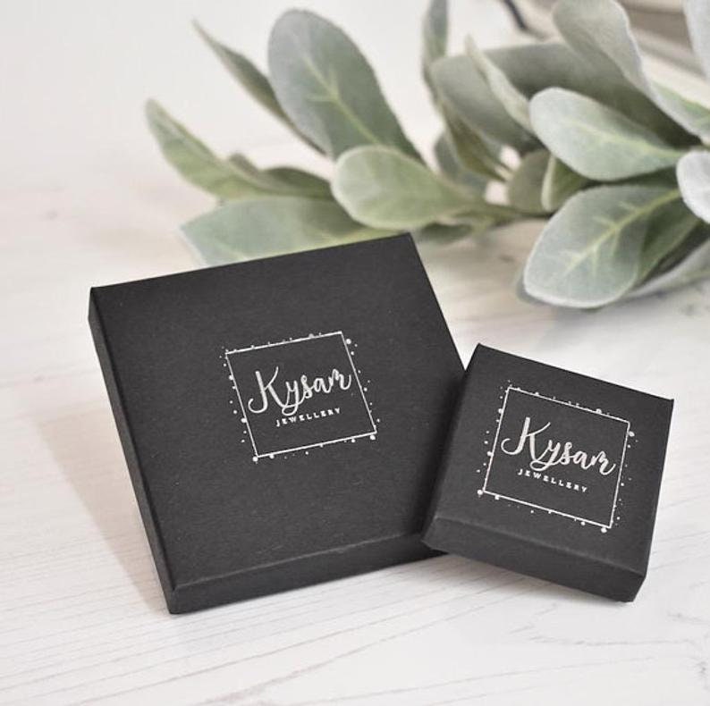 May include: Two black square boxes with the text "Kysar Jewellery" in white lettering. The boxes are on a white wooden surface.