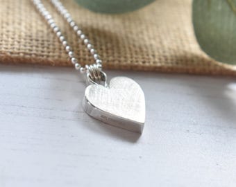 May include: A silver heart pendant on a ball chain necklace. The heart-shaped charm has a brushed metal finish and is suspended from a delicate chain. The necklace is displayed on a neutral-toned surface, with a blurred background.