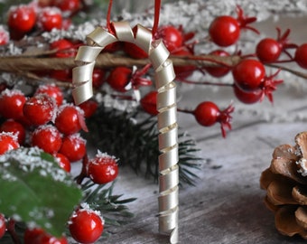 May include: A silver-colored, ribbon-like candy cane Christmas ornament. The ornament is hanging from a red ribbon. The background includes red berries, pine needles, and a pine cone, suggesting a festive holiday theme.