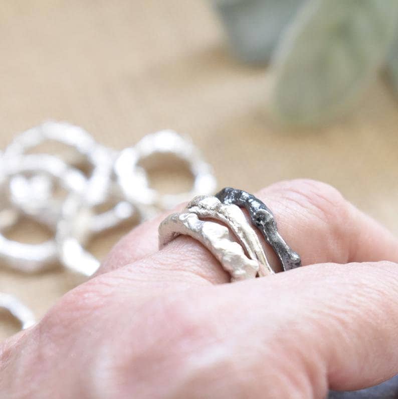 May include: Three silver rings with a textured, organic design. The rings are stacked on a finger, with the top ring being black.