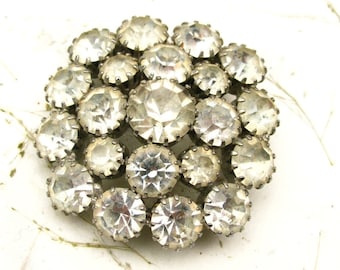 May include: A cluster of clear, faceted rhinestones set in silver metal. The rhinestones are arranged in a circular pattern and have a vintage look.