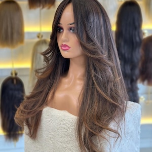 May include: A mannequin head displaying a long, layered wig with brown and auburn highlights. The wig features a natural-looking part and cascading waves. Other wigs are visible in the background, suggesting a hair product display.