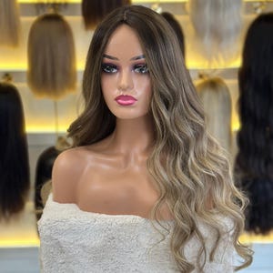 May include: A long, wavy wig featuring a rooted dark brown base that transitions into lighter ash blonde highlights. The hair is styled with a center part and soft, loose waves that cascade down the shoulders, creating a natural, multi-tonal appearance.