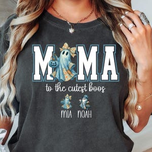 May include: Dark grey t-shirt featuring the word "MAMA" in white block letters with a blue outline. A ghost graphic is in the centre. Below "MAMA" is the phrase "to the cutest boos" and two ghost graphics. The names "MIA NOAH" are printed below.
