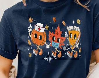 May include: Navy blue t-shirt featuring a whimsical design of three cartoon characters: a pumpkin nurse, a leaf wearing a mask, and a slice of pie with a stethoscope. The design includes fall leaves and a heartbeat line.