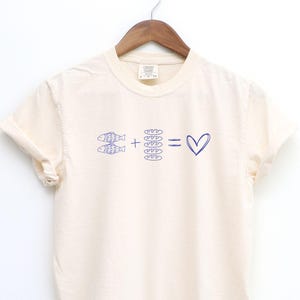 May include: Cream-colored t-shirt with a blue graphic. The graphic shows two fish plus a stack of bread equals a heart. The shirt is hanging on a wooden hanger.