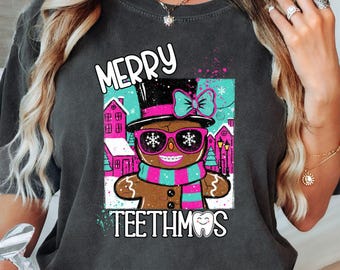 Könnte beinhalten: Dunkelgraues T-Shirt mit einem Lebkuchenmann mit Sonnenbrille, Zylinder und Schal. Der Text "MERRY TEETHMOS" befindet sich über und unter der Grafik. Das Design umfasst rosa, blaue und weiße Akzente mit einem winterlichen Hintergrund.