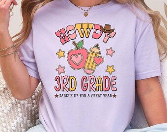 Op de afbeelding: Een lichtpaars T-shirt met de woorden "HOWDY 3RD GRADE" in roze en oranje letters. Het shirt heeft een appel, potlood en sterren afbeelding. De zin "SADDLE UP FOR A GREAT YEAR" staat eronder gedrukt.