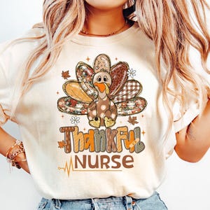 May include: Cream-colored t-shirt featuring a Thanksgiving turkey design with the words "Thankful Nurse." The turkey has patterned feathers in shades of brown, orange, and beige. The text includes a heartbeat line, suggesting a medical theme.