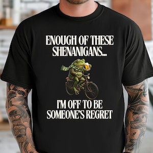 Könnte beinhalten: Schwarzes T-Shirt mit dem Text "ENOUGH OF THESE SHENANIGANS... I'M OFF TO BE SOMEONE'S REGRET." Ein Cartoon-Frosch, der einen Schal trägt und einen Bierkrug hält, fährt Fahrrad. Der Frosch ist grün und der Text ist weiß.