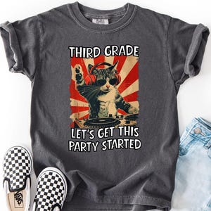 Puede incluir: Una camiseta gris carbón con el gráfico de un gato con auriculares rojos y gafas de sol pinchando en un tocadiscos. El texto dice THIRD GRADE y LET'S GET THIS PARTY STARTED, sobre un fondo de rayos de sol en color rojo y blanco.