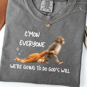 May include: A grey Comfort Colors t-shirt with the text "C'mon Everyone, We're Going to Do God's Will." The shirt features a graphic of a monkey pulling a stuffed animal. A silver necklace with a pendant is on the shirt.