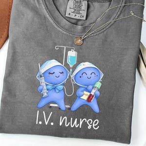 May include: A gray Comfort Colors t-shirt featuring a graphic of two blue cartoon nurses with medical equipment. The text "I.V. nurse" is printed in white below the graphic. A gold necklace with a pendant is draped across the shirt.