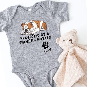 Könnte beinhalten: Ein grauer Baby-Body mit dem Text "PROTECTED BY A SNORING POTATO" und einem Pfotenabdruck. Der Body zeigt eine schlafende Bulldoggen-Illustration in Braun und Weiß. Ein beiger Teddybär und eine Decke liegen neben dem Body.