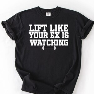 Könnte beinhalten: Schwarzes T-Shirt mit dem weißen Text "LIFT LIKE YOUR EX IS WATCHING" und einer Hantel-Grafik. Das Shirt besteht aus weichem Material und hat einen Rundhalsausschnitt. Ideal für Fitness-Enthusiasten.