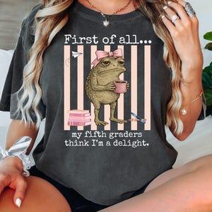 May include: A charcoal gray t-shirt featuring a graphic of a toad wearing a pink bow, holding a pink mug, and standing before vertical pink stripes. The text reads, First of all... my fifth graders think I'm a delight, with books and a pencil.