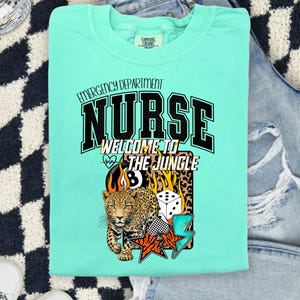 May include: A light teal Comfort Colors t-shirt with the words "Emergency Department Nurse" and "Welcome to the Jungle" in black and white. The graphic includes a leopard, dice, and stars.