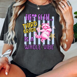 Puede incluir: Una camiseta gris carb&oacute;n de manga corta con el gr&aacute;fico de un mapache rosa sosteniendo una margarita. El texto "FIRST OF ALL... THIRD GRADE IS A WHOLE VIBE" aparece en amarillo y morado sobre un fondo de rayas verticales de color azul claro.
