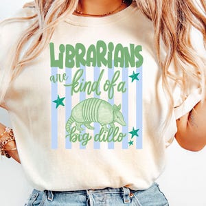 May include: A cream-colored t-shirt featuring a graphic with light blue vertical stripes and the green text "LIBRARIANS are kind of a big dillo". The design includes a green armadillo illustration and several small green stars surrounding the text.