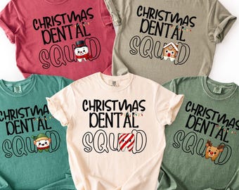 Könnte beinhalten: Fünf T-Shirts in verschiedenen Farben mit dem schwarzen Schriftzug "CHRISTMAS DENTAL SQUAD". Jedes Shirt zeigt ein anderes weihnachtliches Motiv, darunter ein Schneemann, ein Lebkuchenhaus, ein Elf, ein Zuckerstangen-Zahn und ein Rentier.