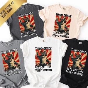 Puede incluir: Cinco camisetas de manga corta en gris oscuro, crema, gris claro, blanco y negro. Cada camiseta presenta un gráfico de un gato con auriculares y gafas de sol en un tocadiscos de DJ, con el texto "THIRD GRADE" y "LET'S GET THIS PARTY STARTED."