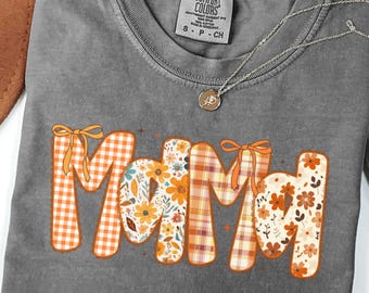 Pode incluir: Uma camiseta cinza Comfort Colors com a palavra "Mamãe" em padrões laranja, florais e xadrez. Um colar de prata com um pingente é visível. A camisa é tamanho S-P-CH.