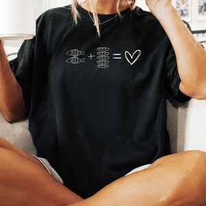 May include: Black t-shirt with a white graphic. The graphic shows two fish plus four bread slices equal a heart symbol. The shirt is a casual style, suitable for everyday wear.