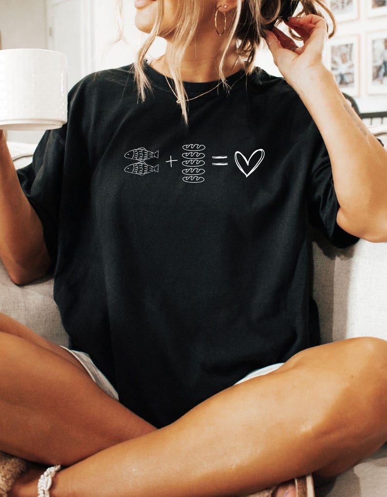 May include: Black t-shirt with a white graphic. The graphic shows two fish plus four bread slices equal a heart symbol. The shirt is a casual style, suitable for everyday wear.
