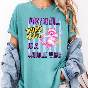 Puede incluir: Una camiseta color verde azulado con un gr&aacute;fico de un mapache rosa sosteniendo una margarita, rodeado de estrellas. El texto dice: FIRST OF ALL... THIRD GRADE IS A WHOLE VIBE. El dise&ntilde;o incluye un fondo de rayas verticales de color azul claro.