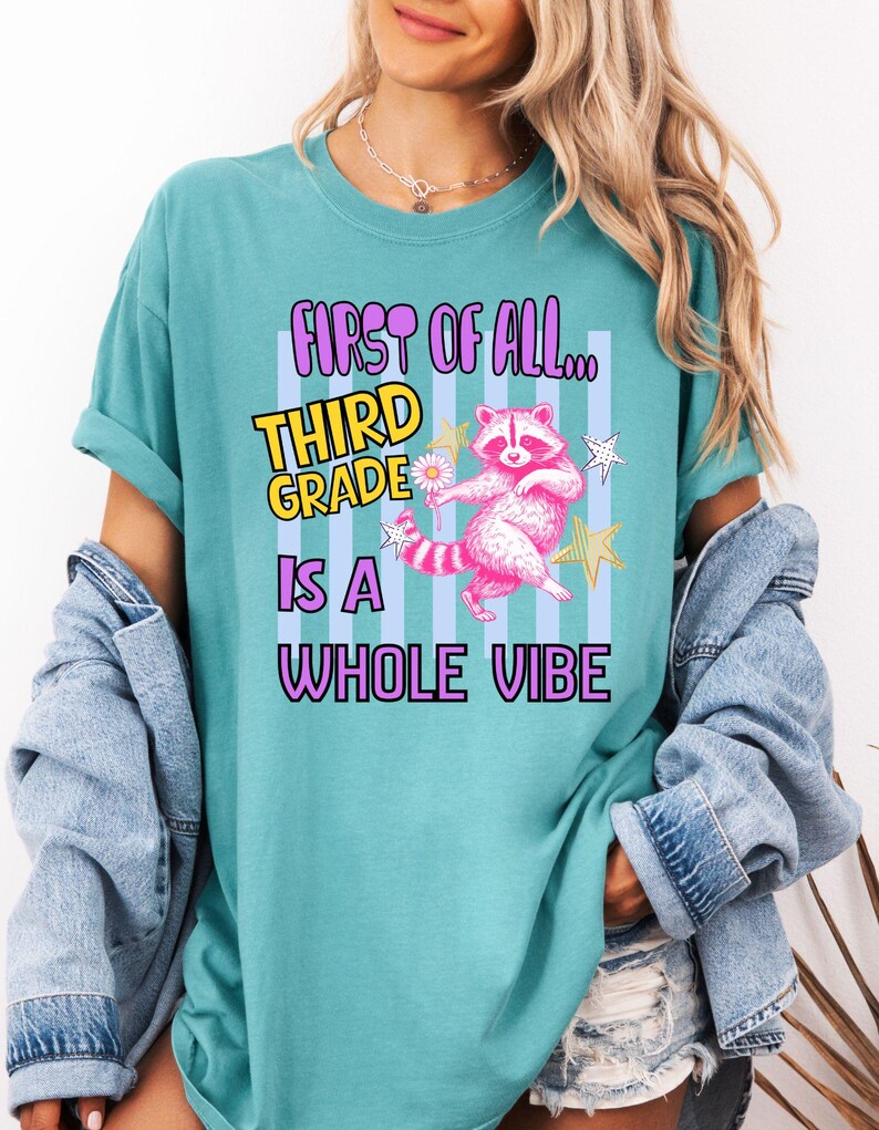 Puede incluir: Una camiseta color verde azulado con un gr&aacute;fico de un mapache rosa sosteniendo una margarita, rodeado de estrellas. El texto dice: FIRST OF ALL... THIRD GRADE IS A WHOLE VIBE. El dise&ntilde;o incluye un fondo de rayas verticales de color azul claro.