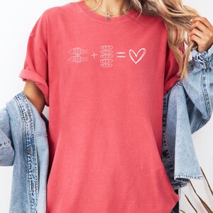 May include: Coral-colored t-shirt with a white graphic. The graphic shows two fish plus four bread rolls equals a heart symbol. The shirt has rolled-up sleeves and is paired with a denim jacket.