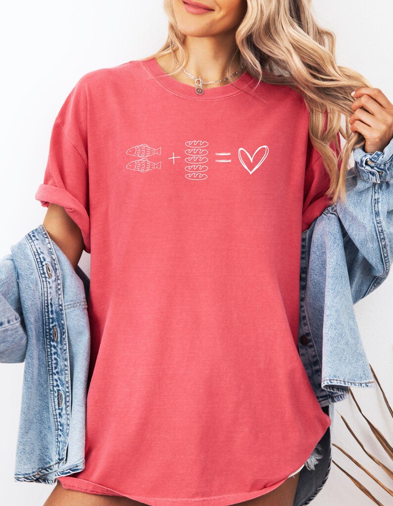 May include: Coral-colored t-shirt with a white graphic. The graphic shows two fish plus four bread rolls equals a heart symbol. The shirt has rolled-up sleeves and is paired with a denim jacket.