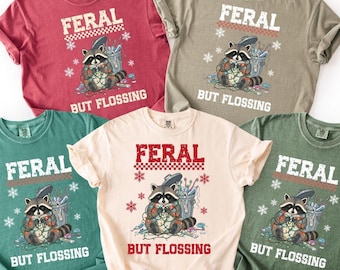 Könnte beinhalten: Fünf verschiedenfarbige T-Shirts mit dem Text "FERAL" über einem Waschbären, der Lichterketten hält, und dem Text "BUT FLOSSING" darunter. Die Shirts sind rot, grün, beige und olivgrün.