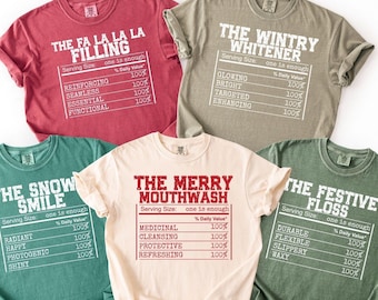 Könnte beinhalten: Fünf T-Shirts in verschiedenen Farben, jeweils mit einem festlichen Text in Weiß, der an Nährwertangaben erinnert. Sätze wie "The Fa La La La Filling" und "The Wintry Whitener" sind zu lesen.