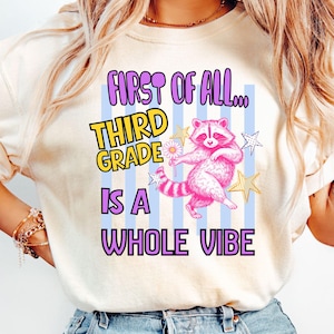 Puede incluir: Una camiseta color crema con un gr&aacute;fico de un mapache rosa sosteniendo una margarita, rodeado de estrellas amarillas y blancas. El texto dice: FIRST OF ALL... THIRD GRADE IS A WHOLE VIBE, sobre un fondo de rayas verticales azul claro.
