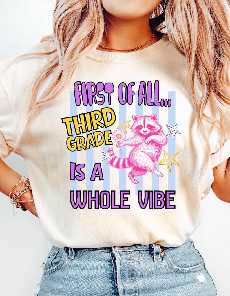 Puede incluir: Una camiseta color crema con un gr&aacute;fico de un mapache rosa sosteniendo una margarita, rodeado de estrellas amarillas y blancas. El texto dice: FIRST OF ALL... THIRD GRADE IS A WHOLE VIBE, sobre un fondo de rayas verticales azul claro.